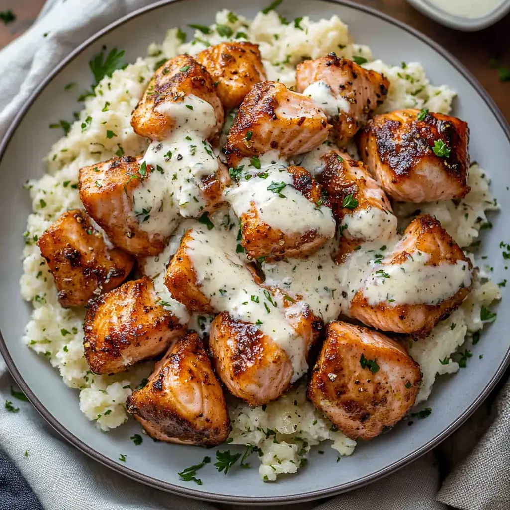 Prato de cubos de salmão grelhados cobertos com molho cremoso, servido sobre uma cama de arroz fofo e decorado com salsa.